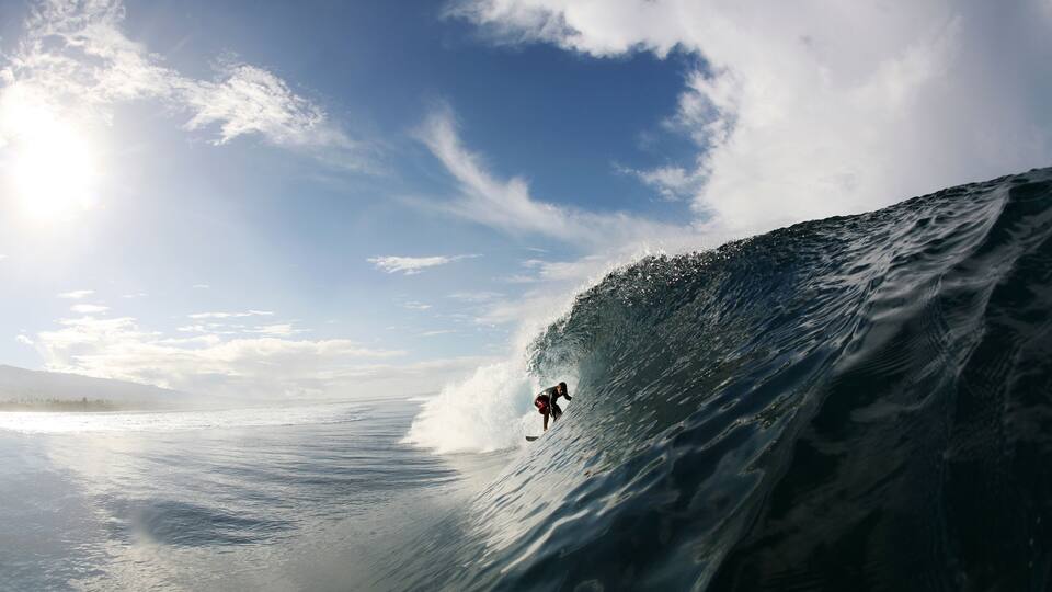 A surfer riding a large wave in the ocean with a clear sky and mountains in the background.