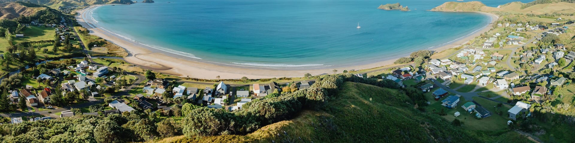 OPITO BAY, COROMANDEL PENINSULA, NEW ZEALAND, Panoramic view of a coastal town nestled beside a beautiful beach. Houses line the shore, with a sailboat visible on the water. Tranquil seaside scene.