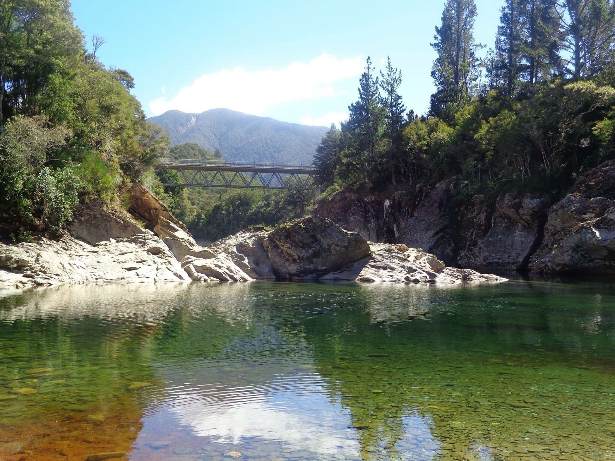 A river with clear water, surrounded by rocky terrain and dense forest. A bridge spans the river in the background.