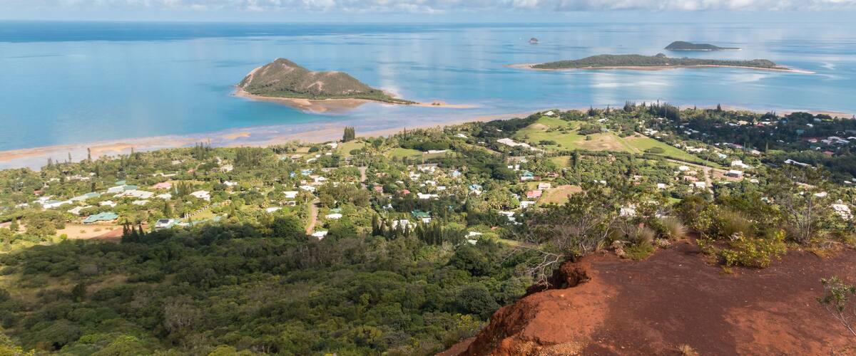 A coastal town with a clear view of the ocean, green hills, and a prominent red rock formation.