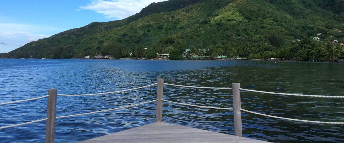A dock with a rope barrier leading to a calm lake, surrounded by lush green hills and a clear blue sky.