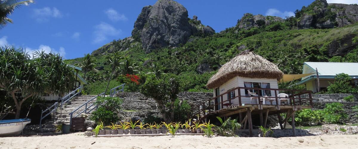 A beach with a thatched-roof hut, palm trees, and a rocky mountain in the background.