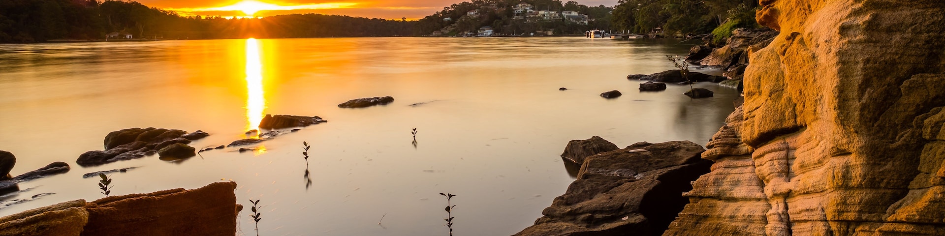 Um pôr do sol sobre um rio calmo, com margens rochosas e edifícios ao longe.