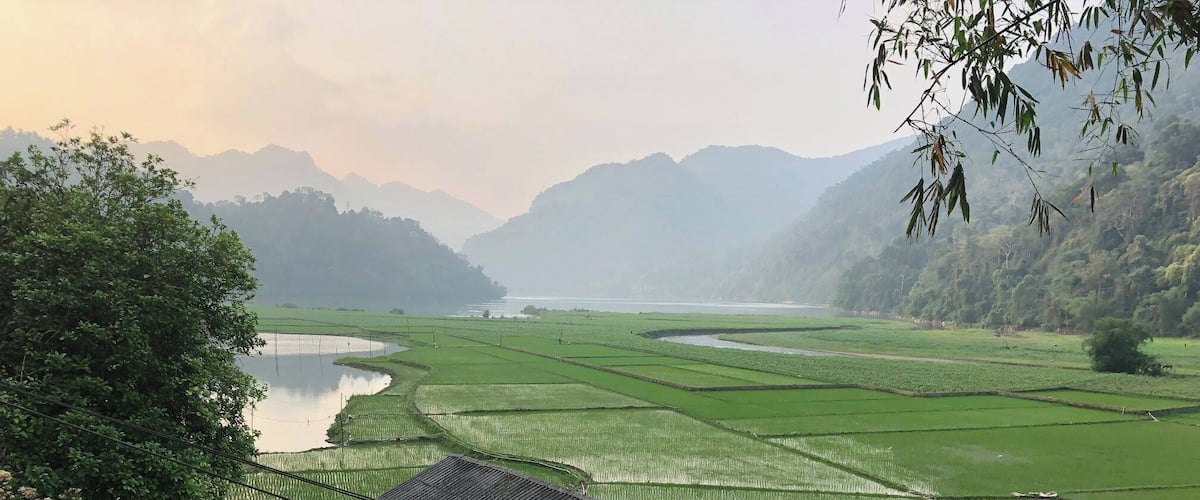 Paisaje rural con campos de arroz en terrazas, una pequeña casa con techo de láminas metálicas y un río al fondo.