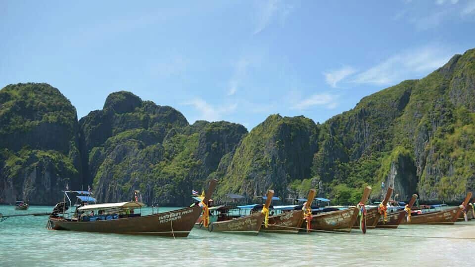 A row of traditional boats docked at a beach with lush green mountains in the background.