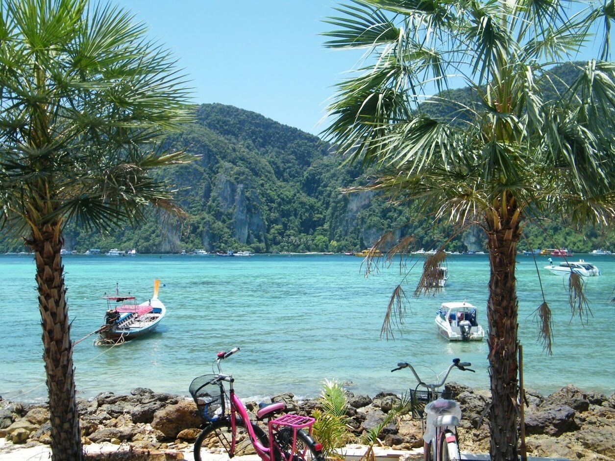 Een kustlandschap met heldere turquoise zee, een rotsachtige oever, palmbomen en verschillende boten die in het water liggen aangemeerd.