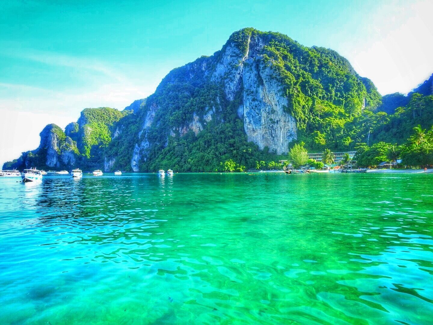 Helder turquoise water met boten, een hoge klif en weelderige begroeiing.