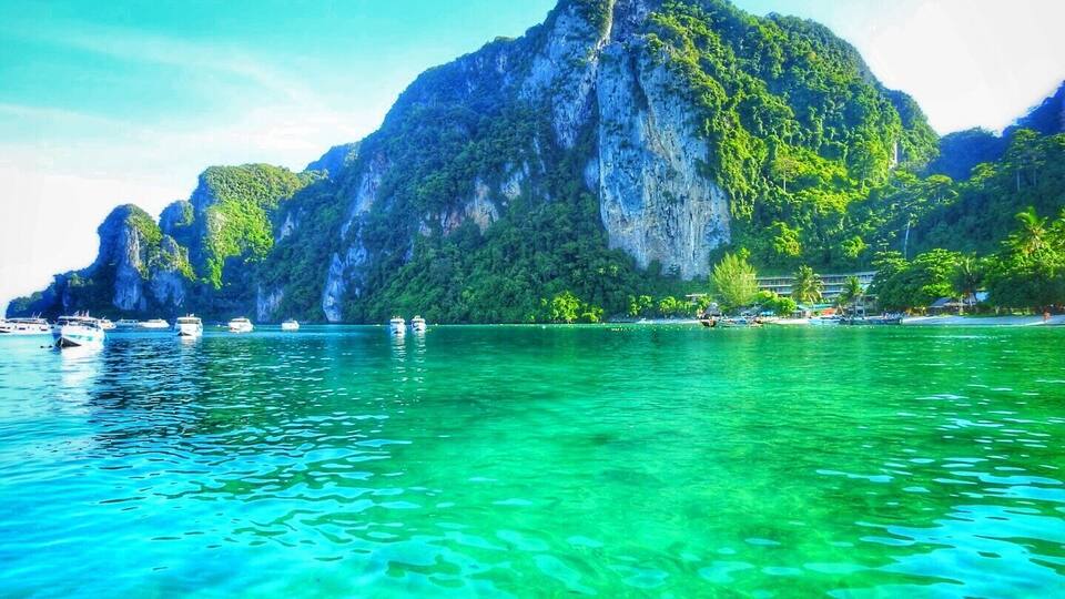 Une mer turquoise aux eaux limpides, parsemée de bateaux, une grande falaise et une végétation luxuriante.