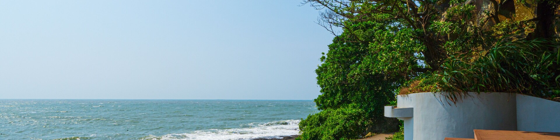A coastal walkway with a rocky shoreline, a clear blue sky, and lush greenery.