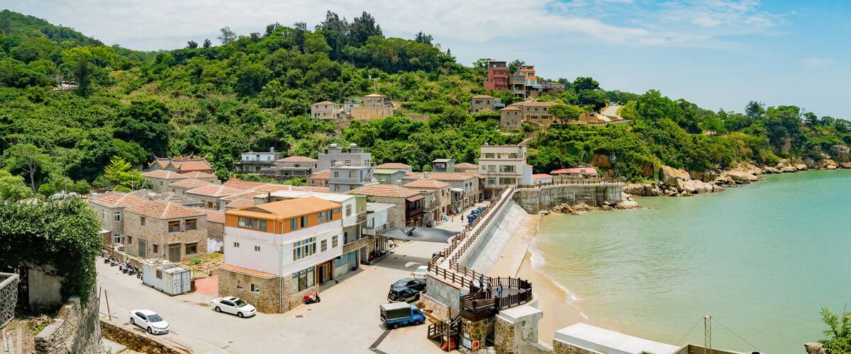 A coastal town with terracotta-roofed buildings, a beach, and a hillside with dense greenery.
