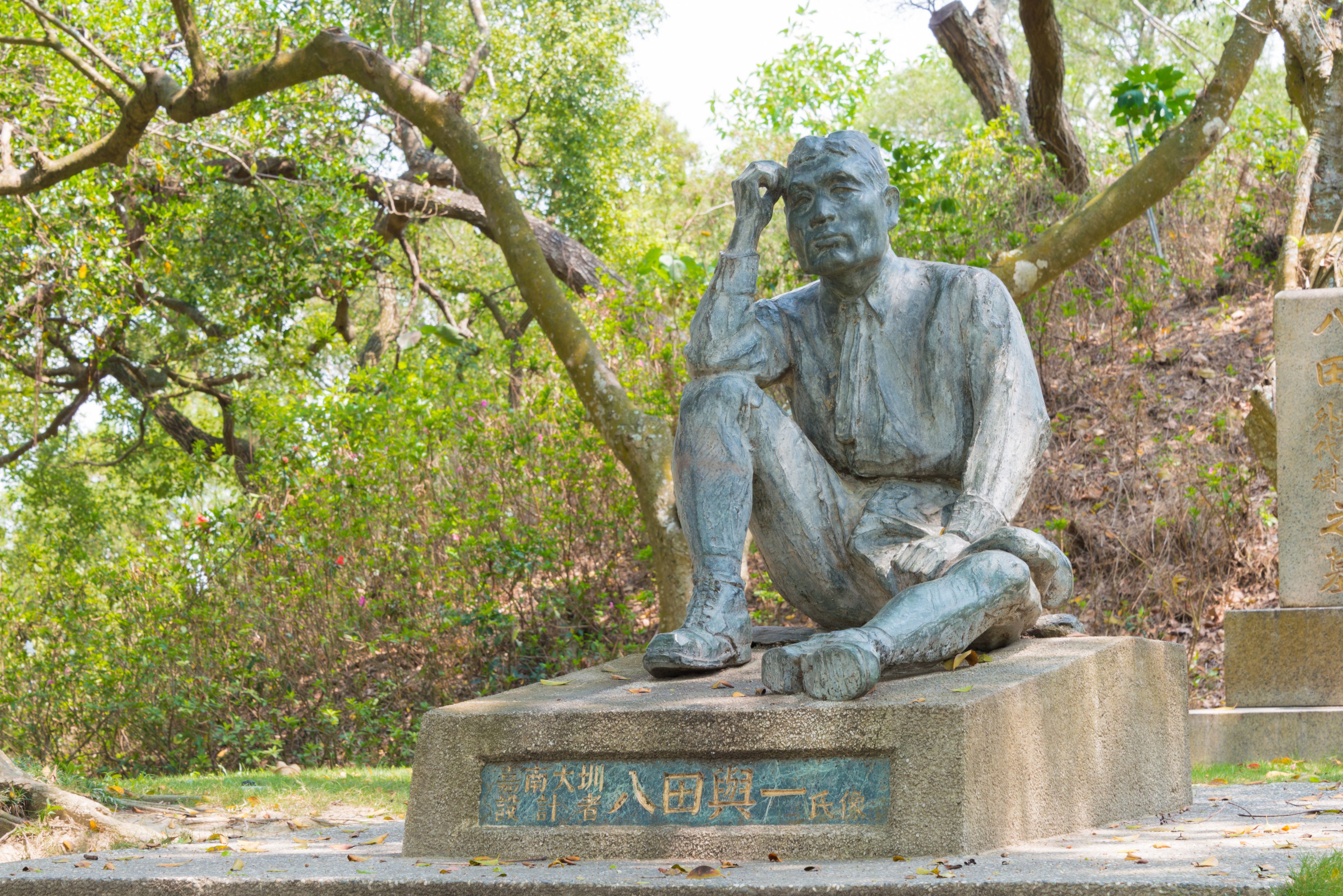 A statue of a seated man with a contemplative pose, surrounded by trees and foliage.