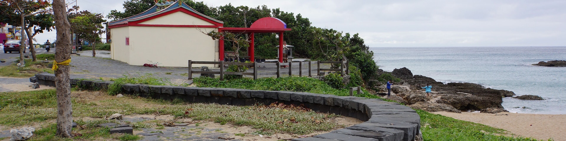 A coastal walkway with a stone wall, a small building with a red roof, and a beach with rocks.