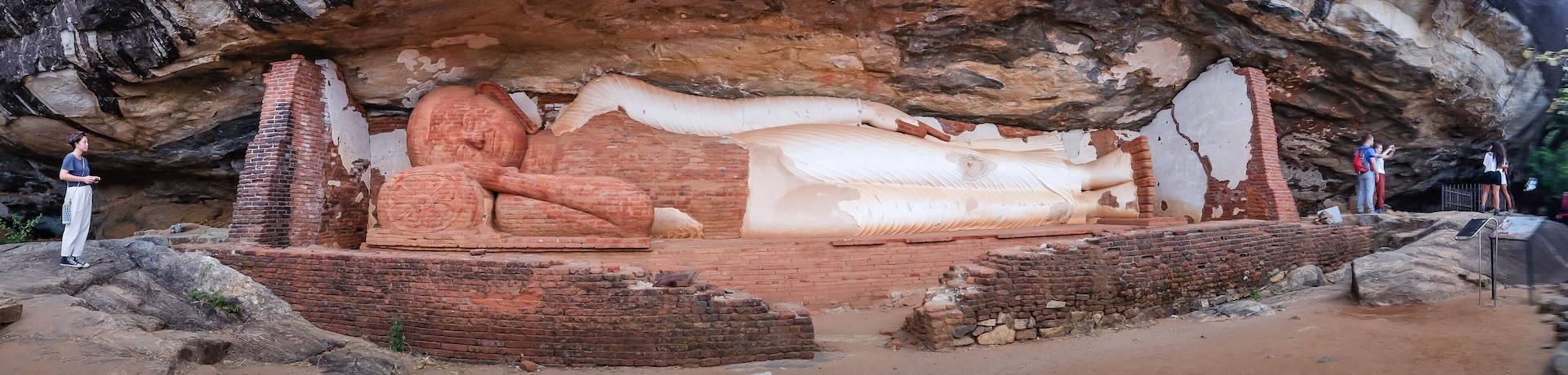 Una gran estatua tallada en roca que representa una figura reclinada, rodeada de construcciones de ladrillo y ubicada frente a un risco rocoso.