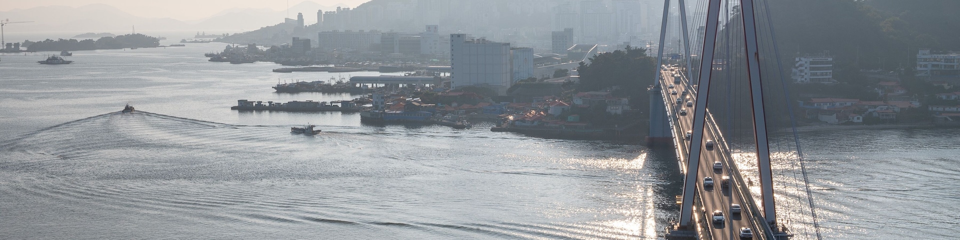 Un puente colgante sobre un cuerpo de agua, con barcos, el perfil de una ciudad y montañas al fondo.