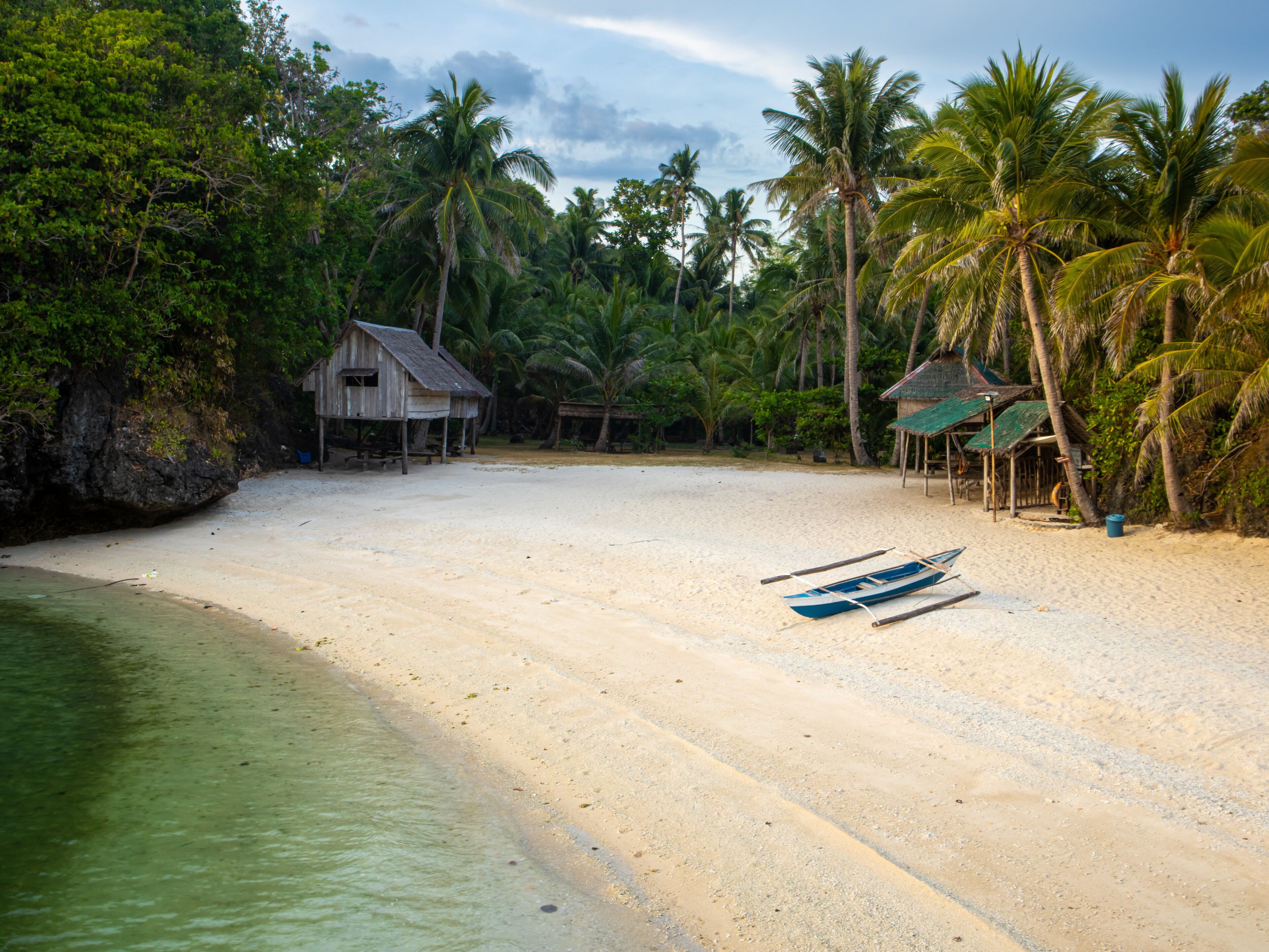 A beach with a wooden hut, palm trees, and a boat on the sand.