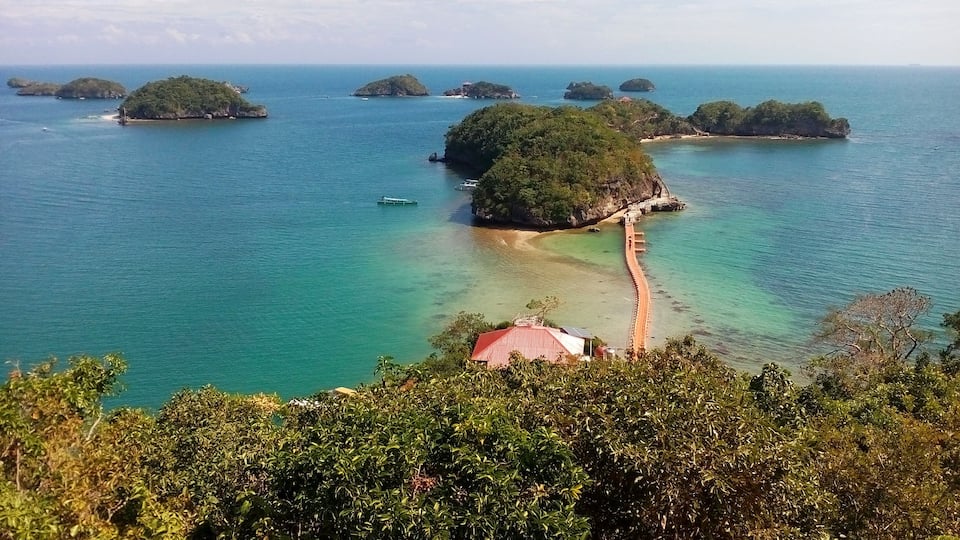 A coastal landscape with several small islands surrounded by turquoise waters. A red-roofed building is visible on the shore, and a white boat is anchored nearby.