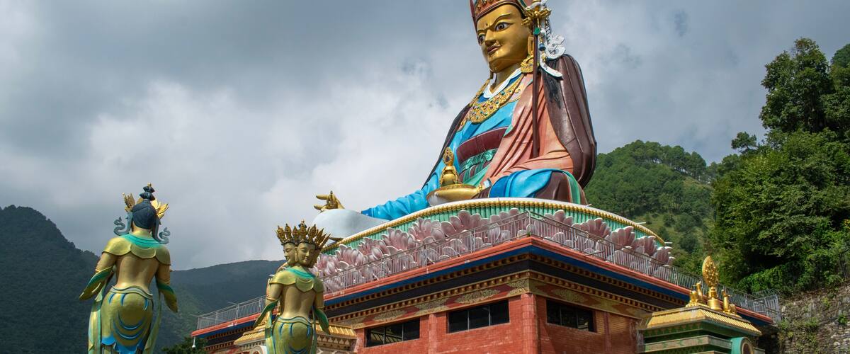 A large statue of a deity with a blue robe and golden jewelry, set against a mountainous backdrop.