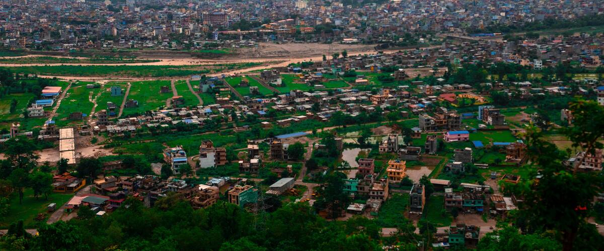 A sprawling cityscape with dense urban development, green spaces, and a distant mountain range.
