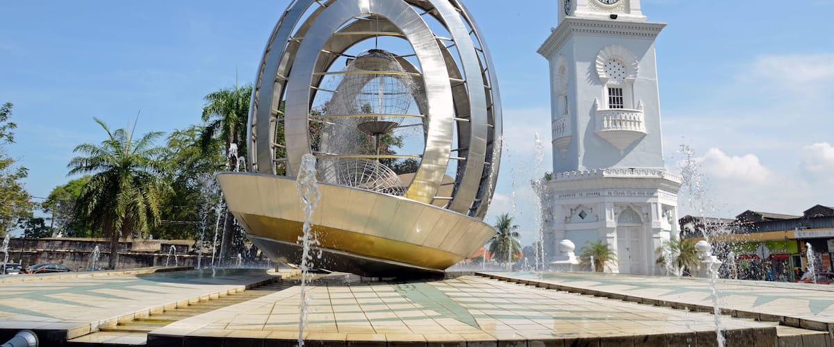 A modern fountain with a large circular structure, a clock tower, and palm trees in the background.