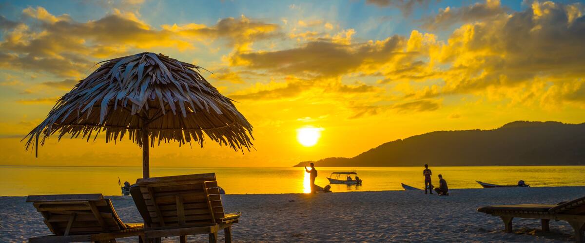 A beach at sunset with a thatched umbrella, wooden lounge chairs, and a calm sea.