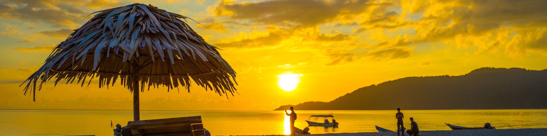 A beach at sunset with a thatched umbrella, wooden lounge chairs, and a calm sea.