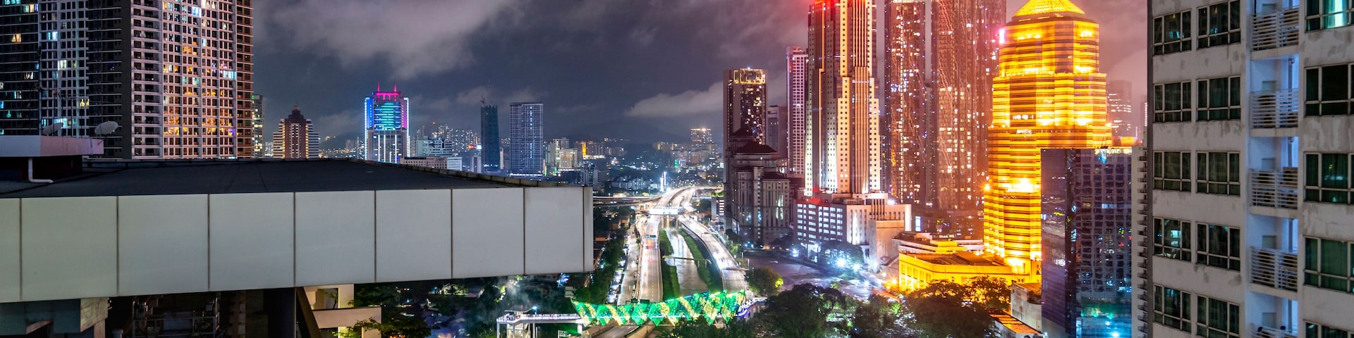 Un paisaje urbano nocturno con rascacielos iluminados, una autopista y zonas verdes circundantes.
