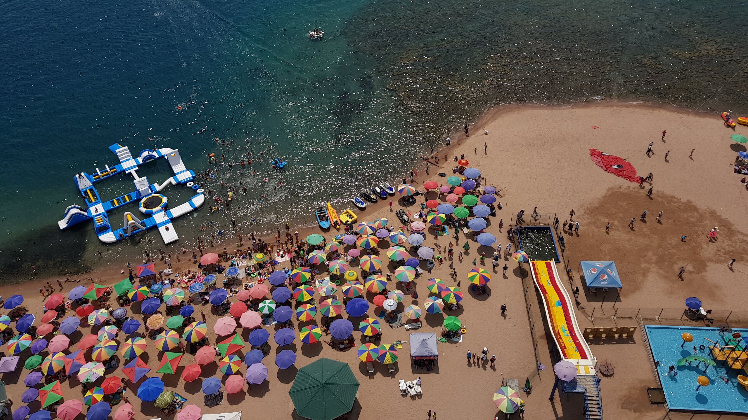 An aerial view of a crowded beach with inflatable structures, colorful umbrellas, and a water slide.