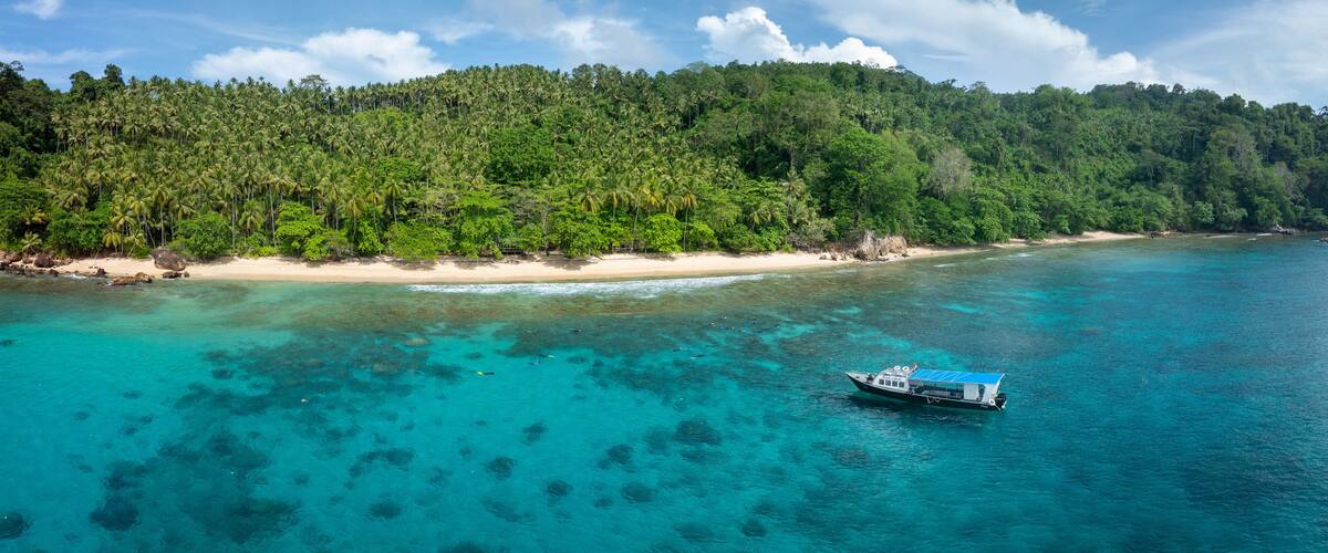 A boat on turquoise waters, a sandy beach, and a forested hill.