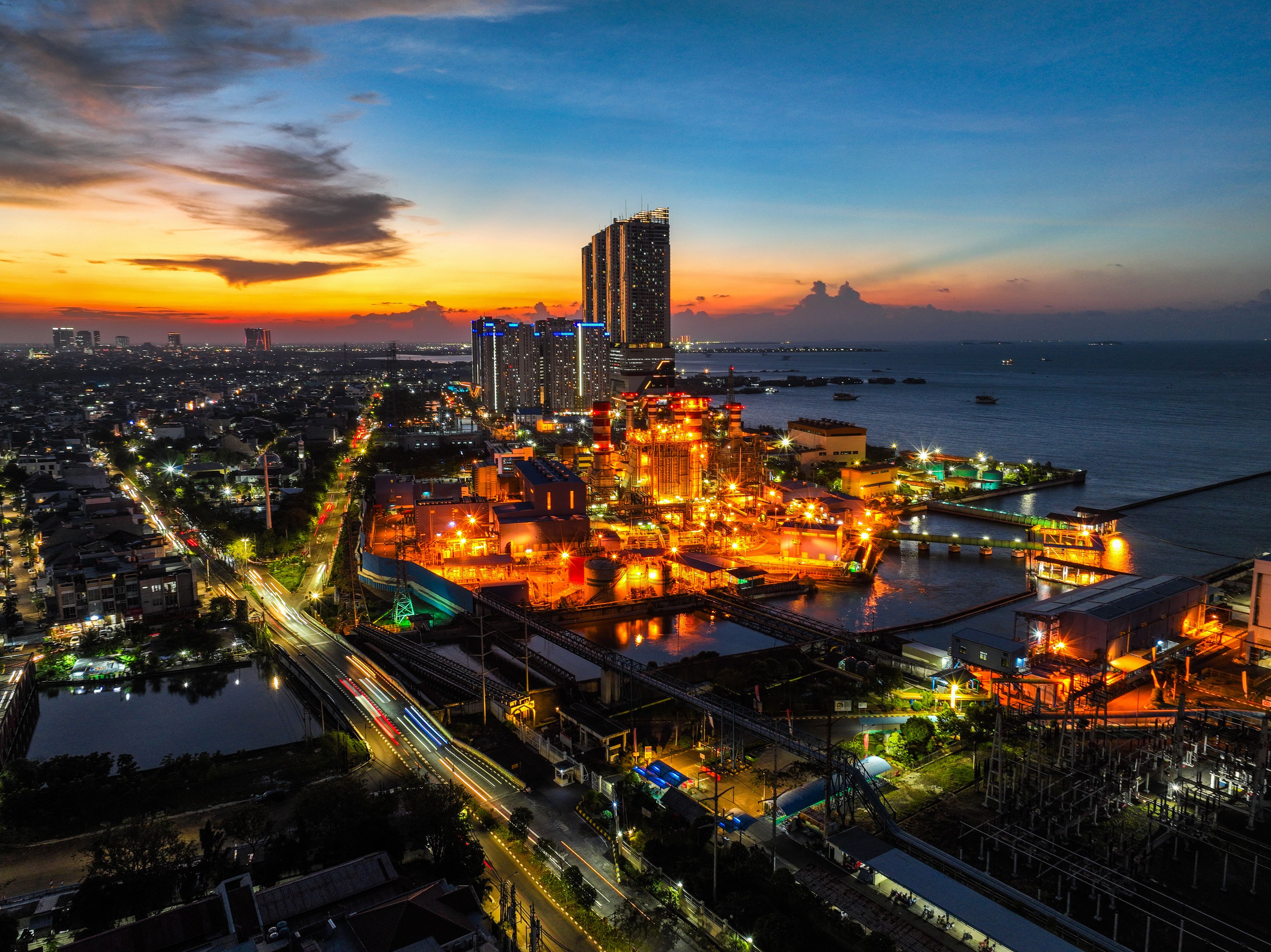 Kecamatan Penjaringan, Daerah Khusus Ibukota, Indonesia - 26 April 2025: Aerial view of the illuminated PLTU Pluit power plant at sunset, with industrial structures and city lights along the coast.