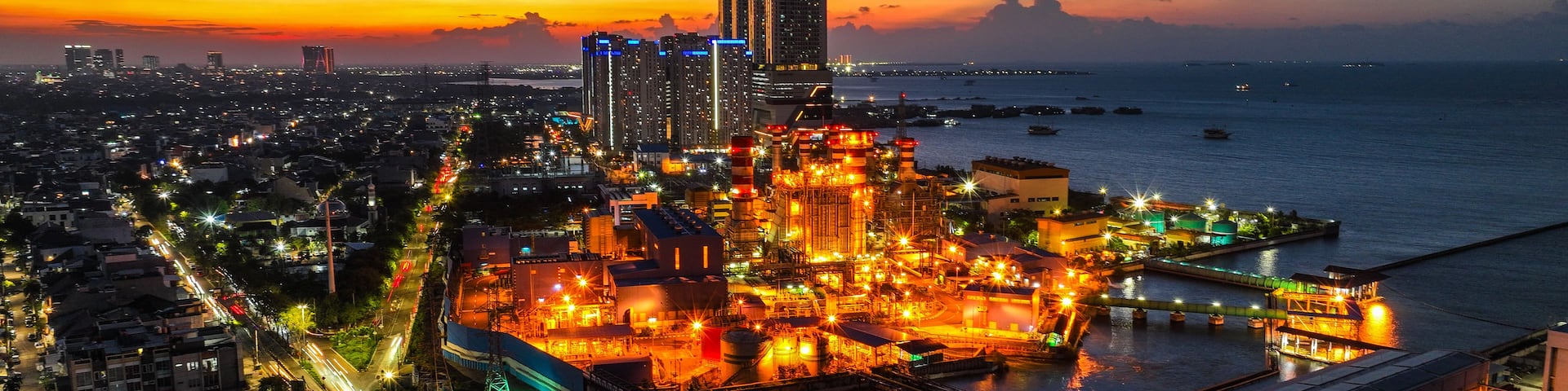 A cityscape at dusk with illuminated industrial areas and a sunset over the water.