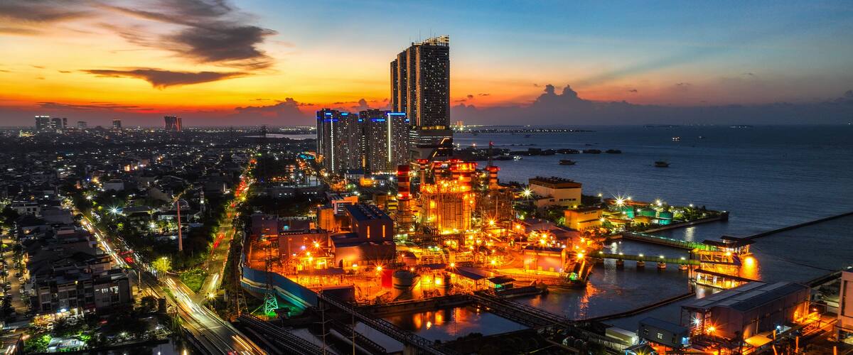 A cityscape at dusk with illuminated industrial areas and a sunset over the water.