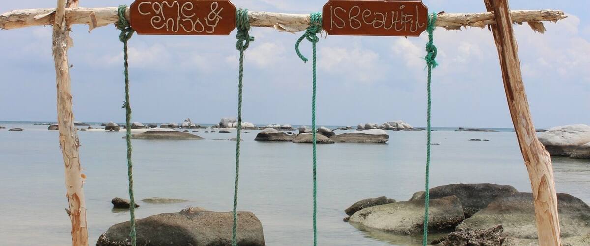 A wooden swing set with signs saying "Come & Relax" and "IS Beautiful" on a sandy beach with calm water and rocks in the background.