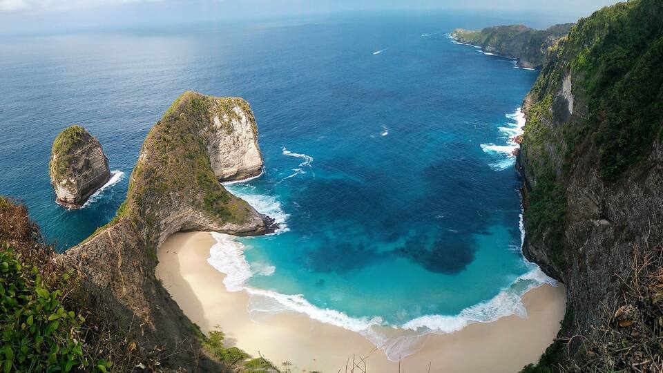 A coastal landscape with a sandy beach, clear blue waters, and rocky cliffs.