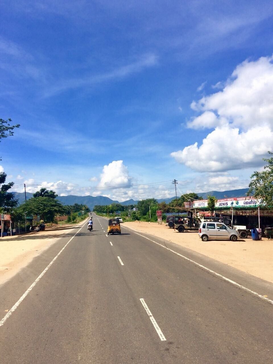 A road with vehicles, roadside shops, and mountains in the background.