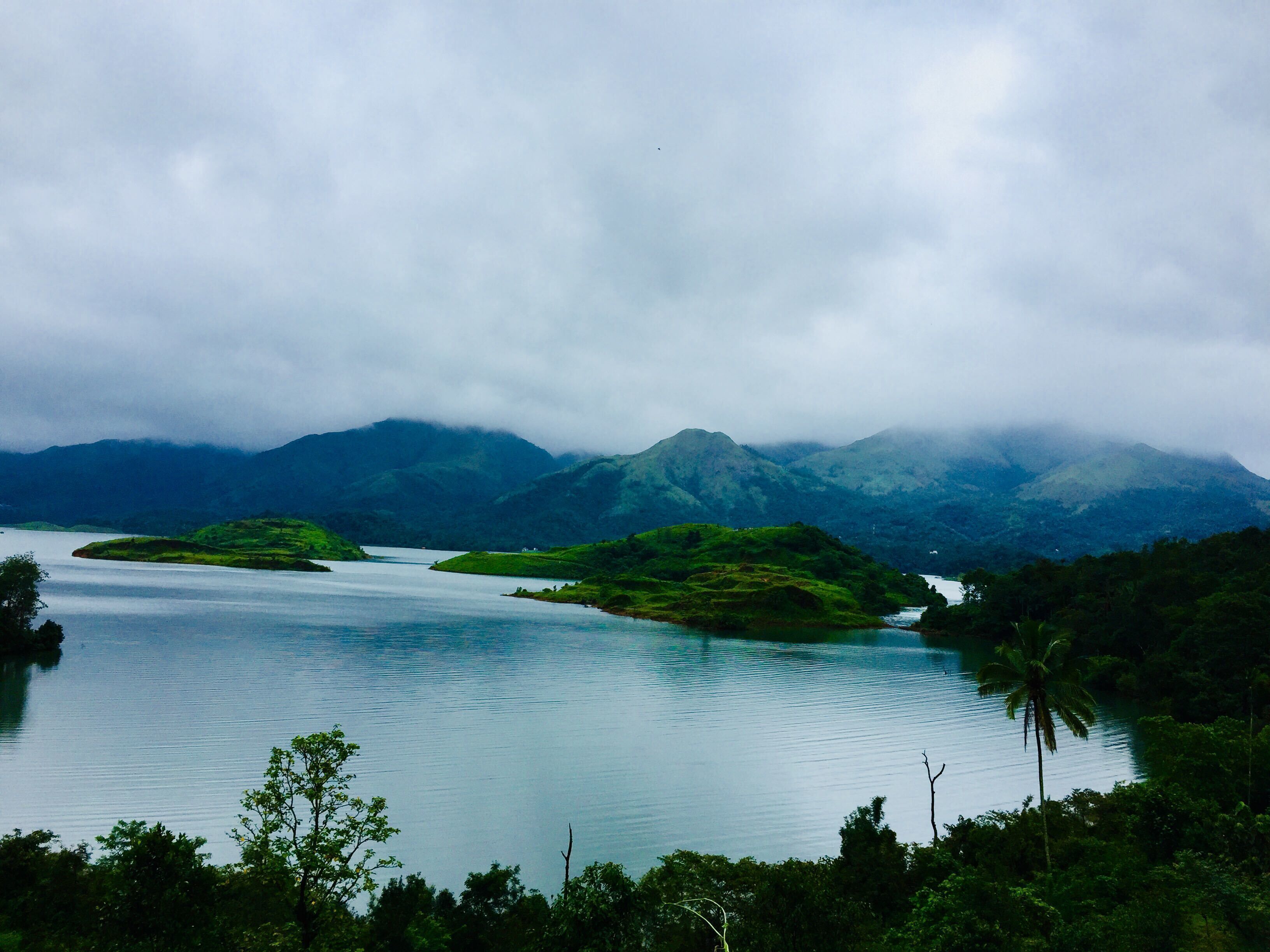 A calm lake surrounded by lush green hills and mountains under a cloudy sky.