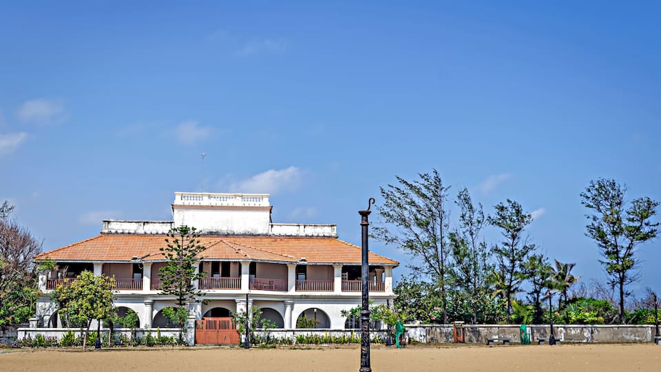 A colonial-style building with a tiled roof and arched windows, surrounded by trees and a sandy area.