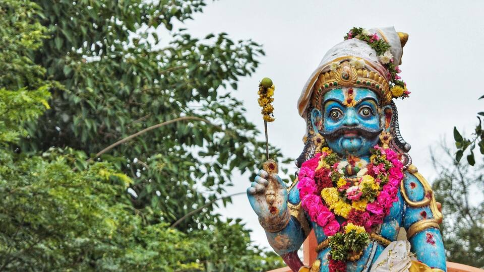 A colorful statue with a blue face, holding a flower and a staff, surrounded by greenery.