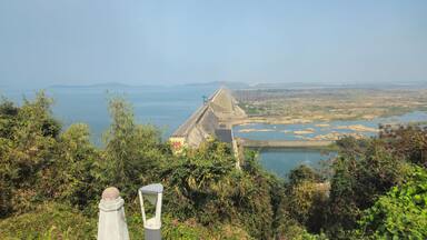 Uma grande barragem com vertedouro, cercada por vegetação exuberante e vista para um rio e montanhas distantes.