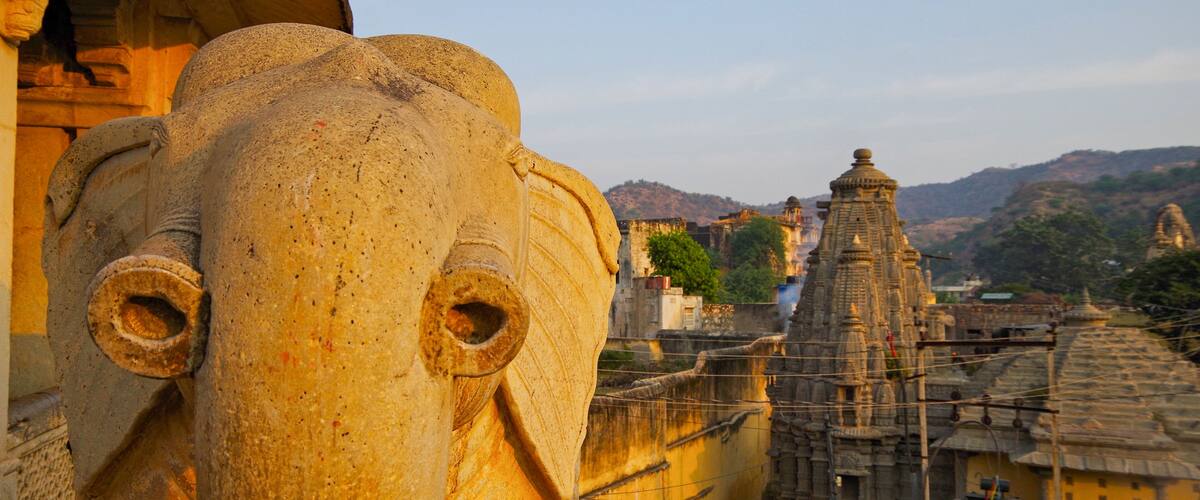 A large stone elephant head sculpture with two tusks, set against a backdrop of a temple and surrounding hills.
