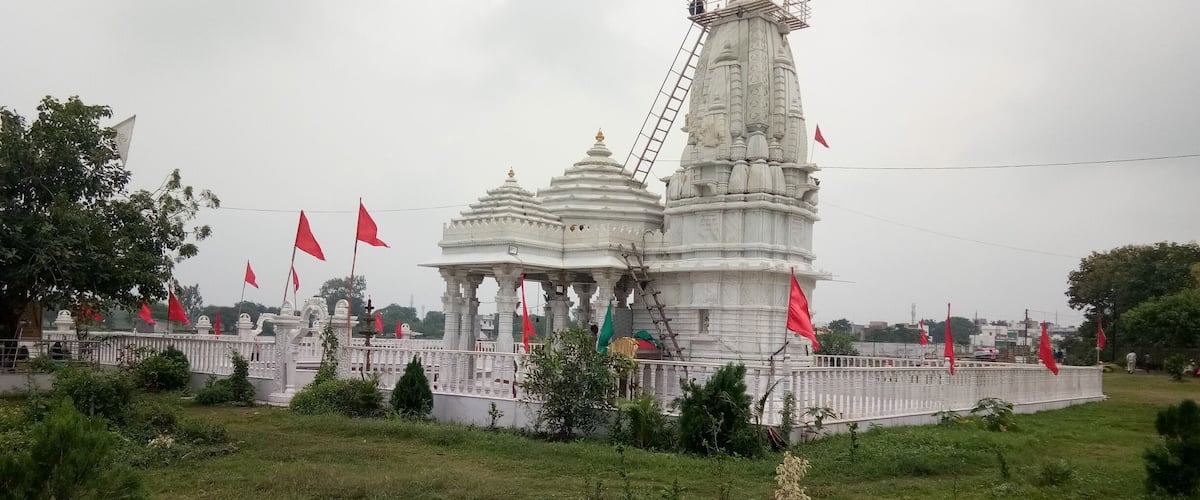 A temple with a central tower and multiple smaller structures, surrounded by a white fence and red flags.