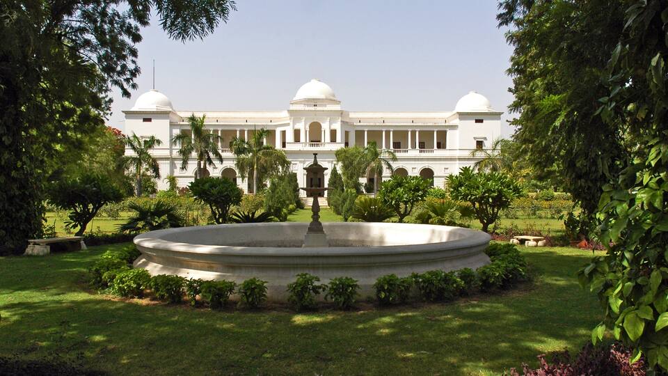 A large white building with domes, surrounded by a well-maintained garden with a central fountain.