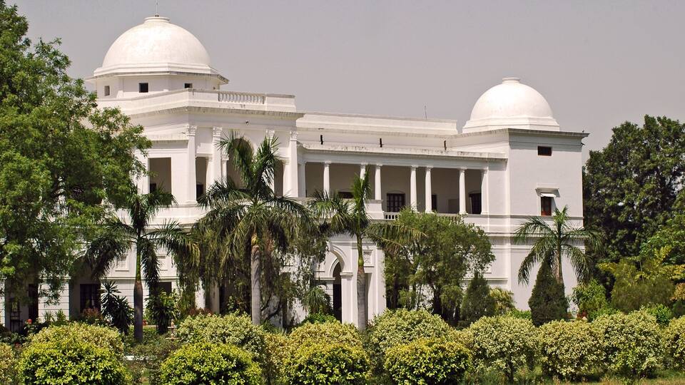 A large white building with two domes, surrounded by lush greenery and palm trees.
