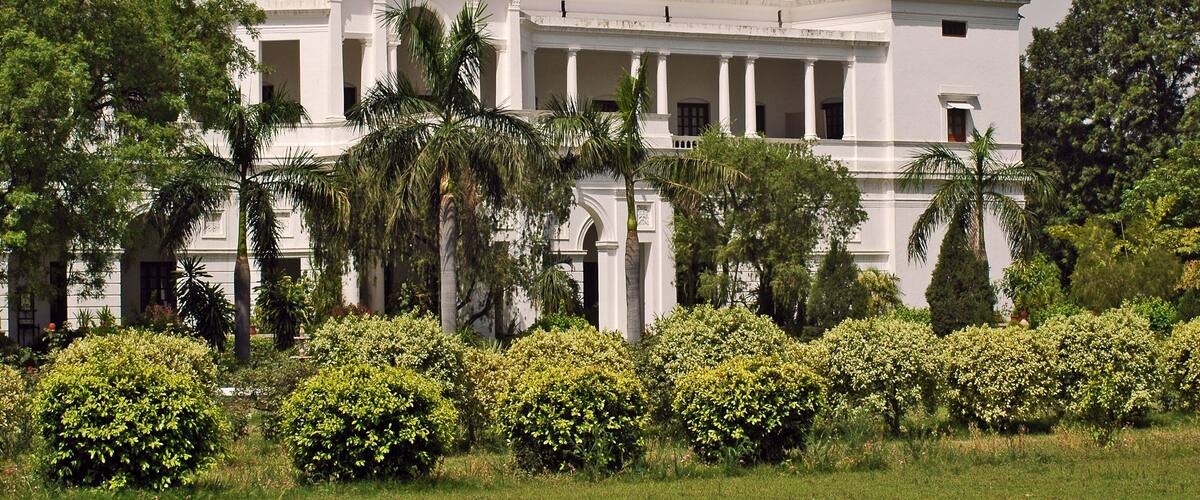 A large white building with two domes, surrounded by lush greenery and palm trees.