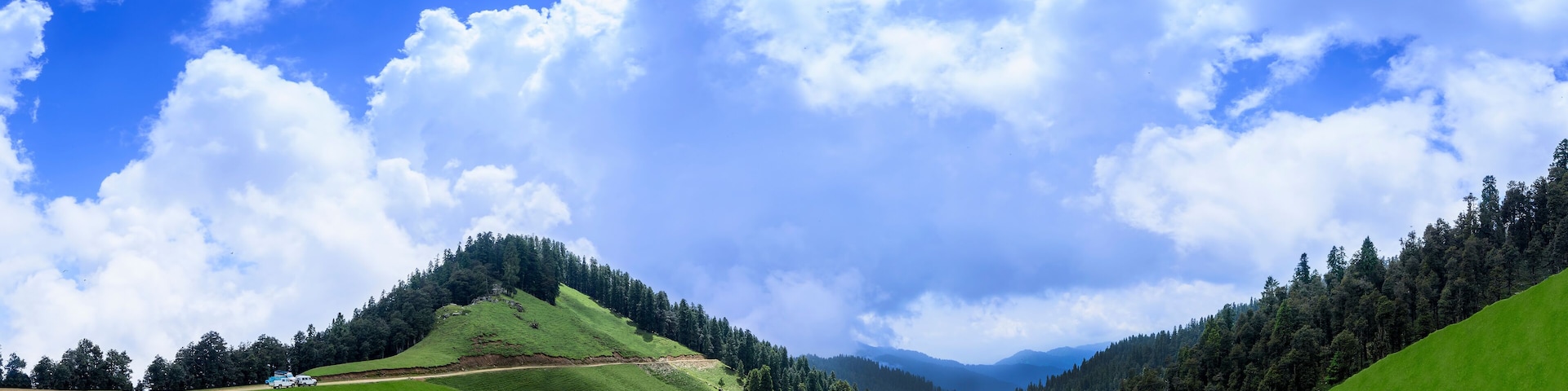 Un valle verde con una carretera, un bosque denso y montañas distantes bajo un cielo azul con nubes.