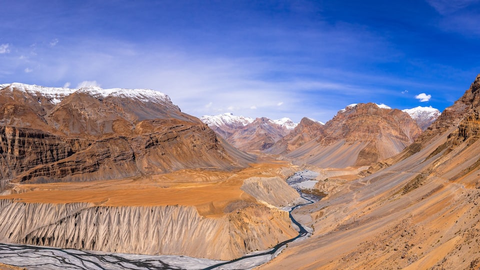 A mountainous landscape with a river flowing through a valley, snow-capped peaks, and a clear blue sky.