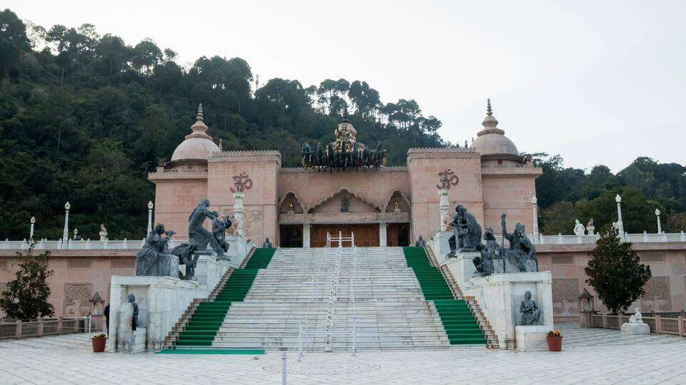 A temple with a staircase leading up to a central entrance, surrounded by statues and a forested hill in the background.