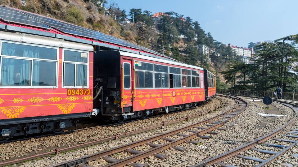 A red and yellow train with the number 094372 on its side, traveling on a curved track surrounded by rocky terrain and trees.