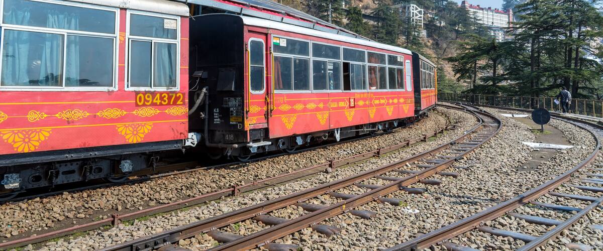 A red and yellow train with the number 094372 on its side, traveling on a curved track surrounded by rocky terrain and trees.