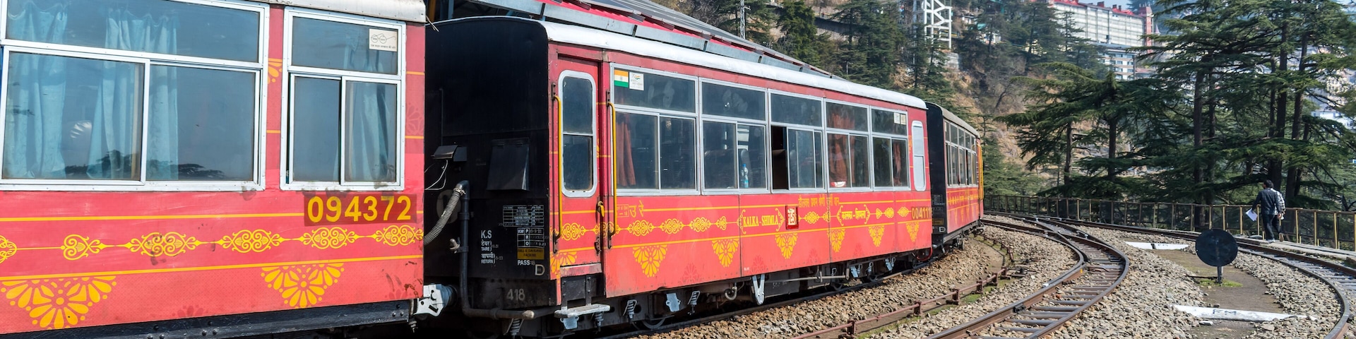 A red and yellow train with the number 094372 on its side, traveling on a curved track surrounded by rocky terrain and trees.