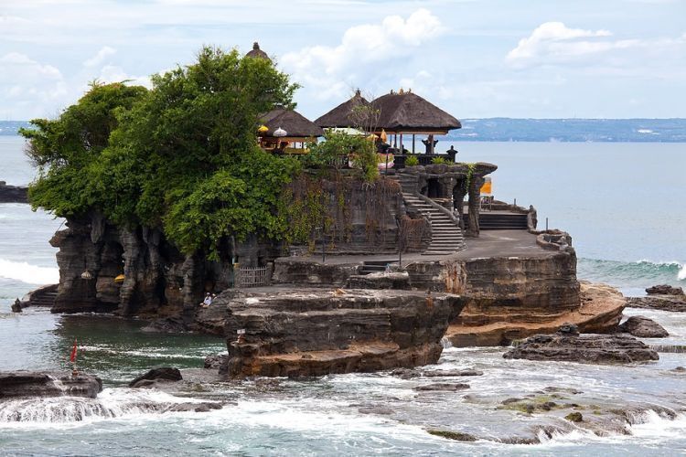 A stone structure with a thatched roof and stairs leading up to a platform, surrounded by lush greenery and overlooking a body of water.